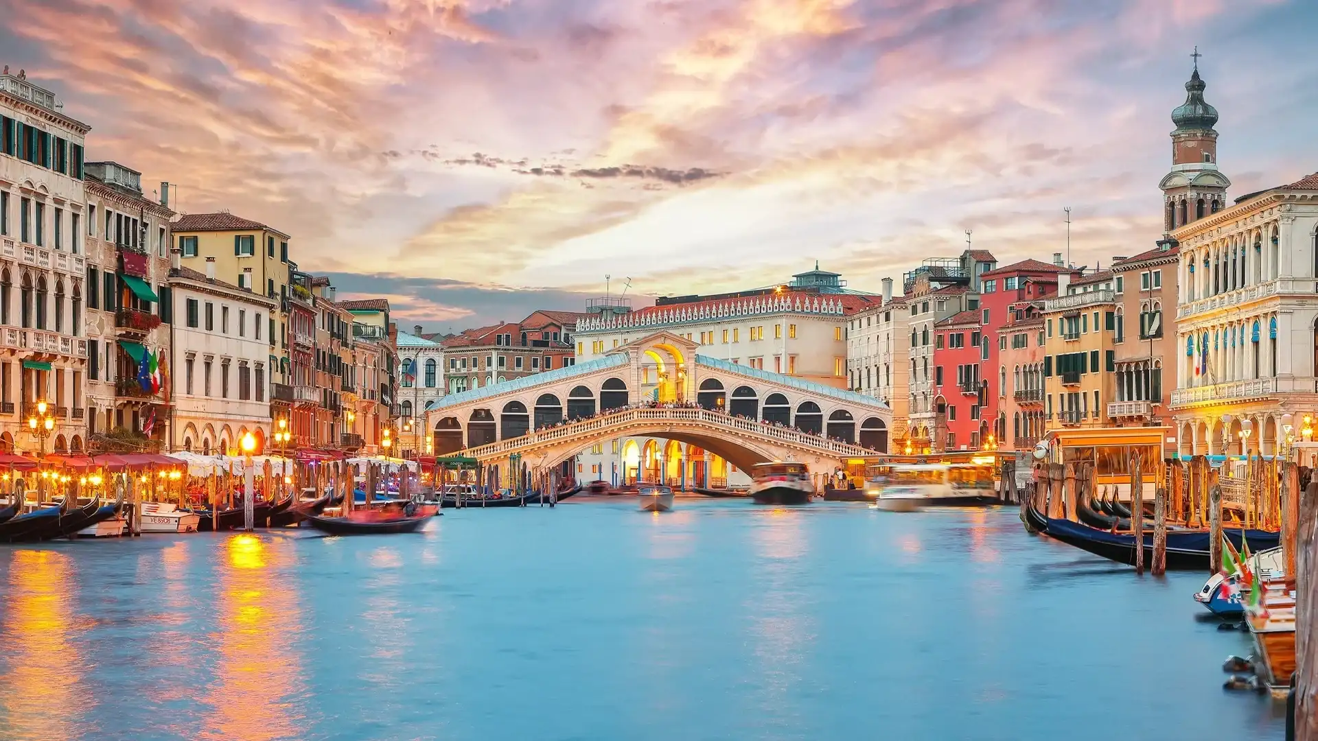 View of the Grand Canal in Venice, Italy, featuring the Rialto Bridge at sunset, surrounded by historic buildings and gondolas on the water
