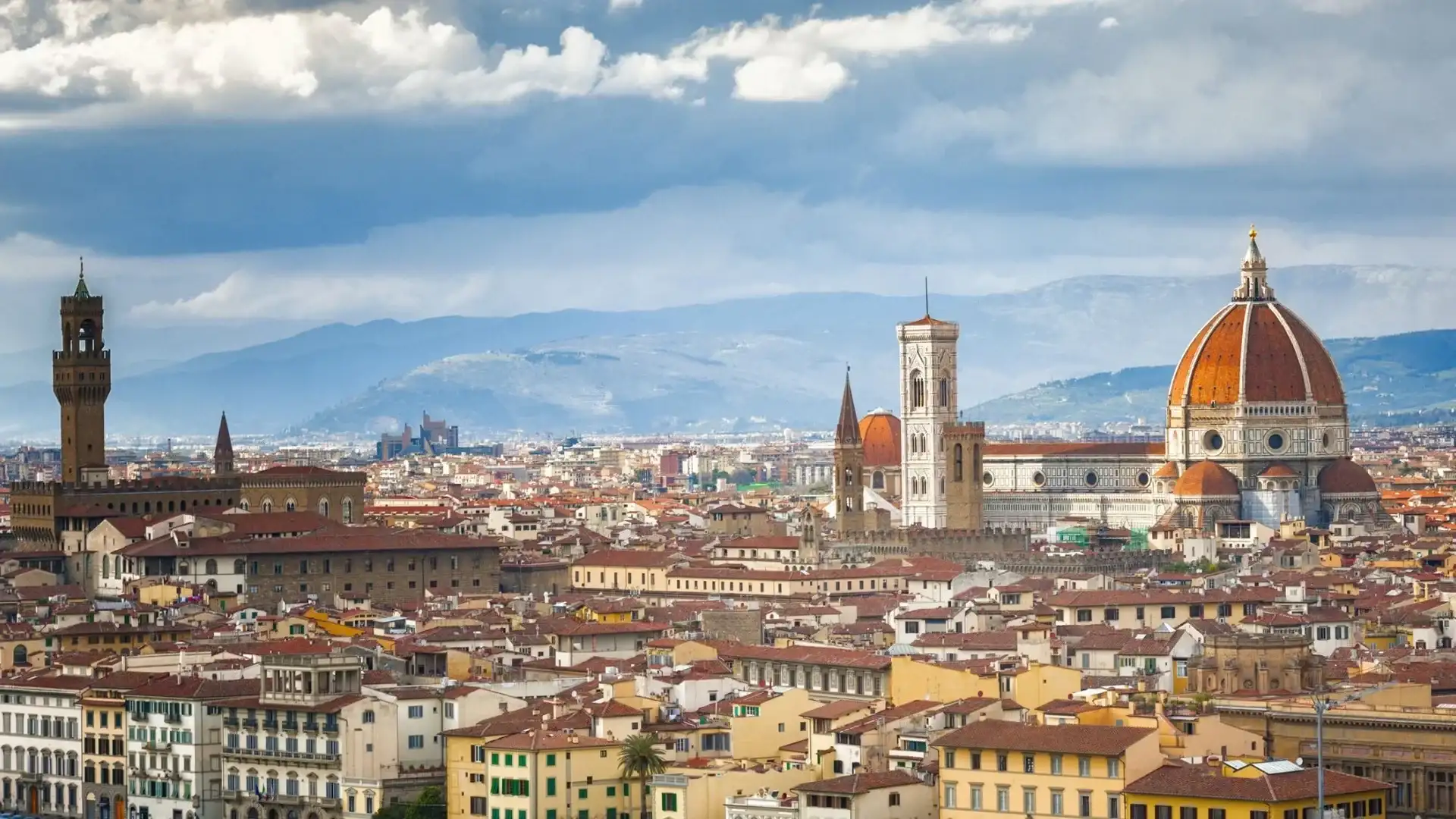 A panoramic view of Florence, showcasing the iconic Duomo, the Palazzo Vecchio, and the Tuscan hills under a cloudy sky.
