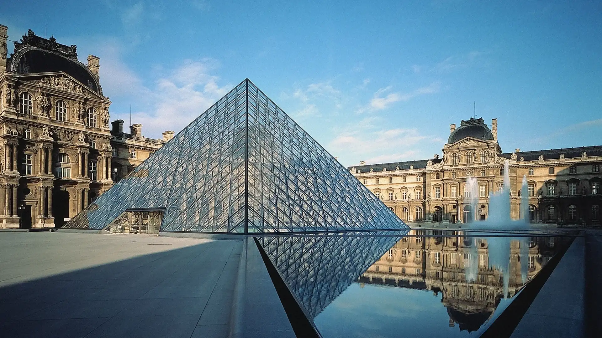 The Louvre Museum features a glass pyramid entrance, reflecting the historic architecture and fountain in the surrounding courtyard.