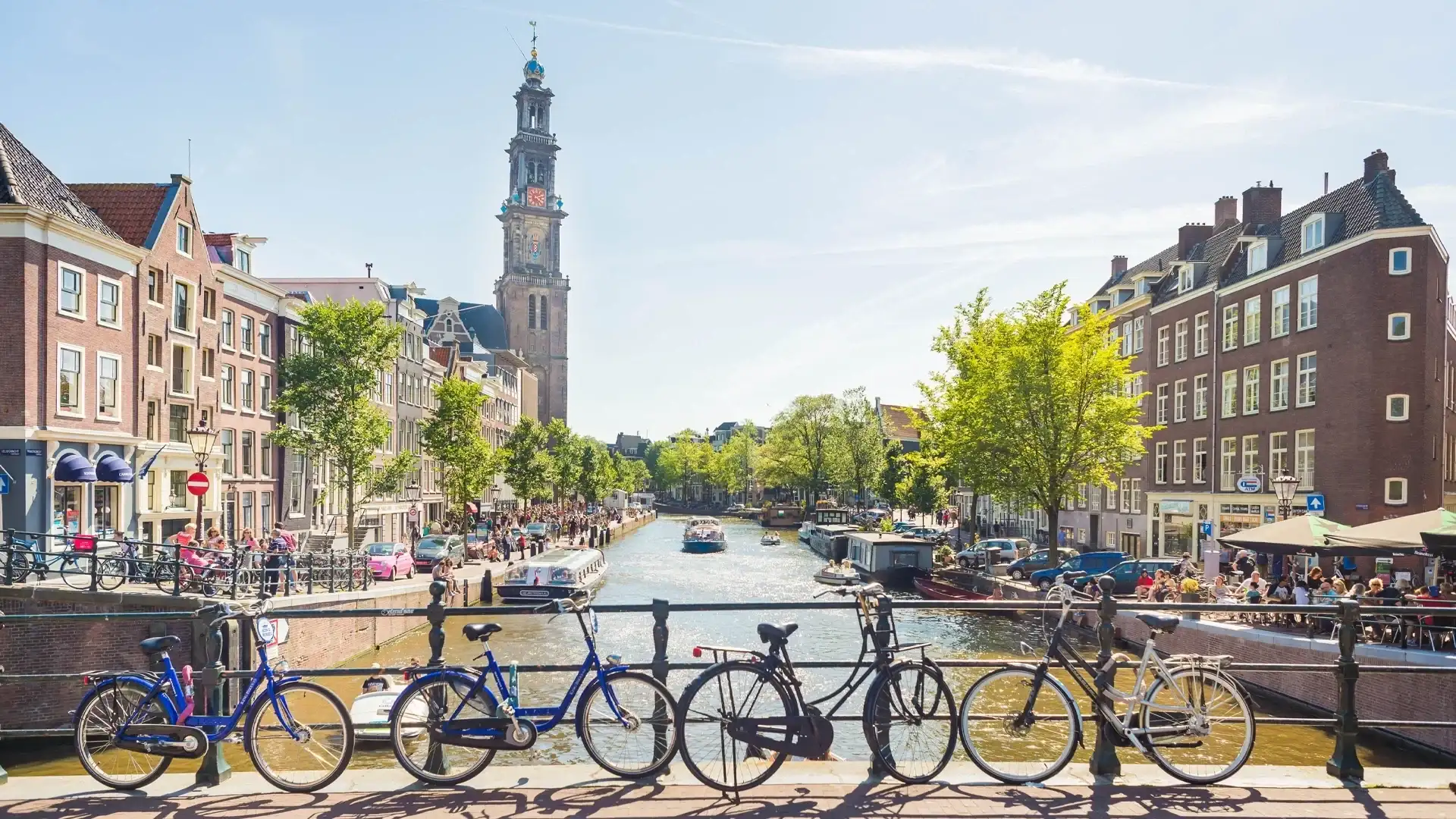 A sunny Amsterdam scene featuring bicycles on a bridge, a canal with boats, and the iconic Westerkerk tower in the background.