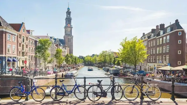 A sunny Amsterdam scene featuring bicycles on a bridge, a canal with boats, and the iconic Westerkerk tower in the background.