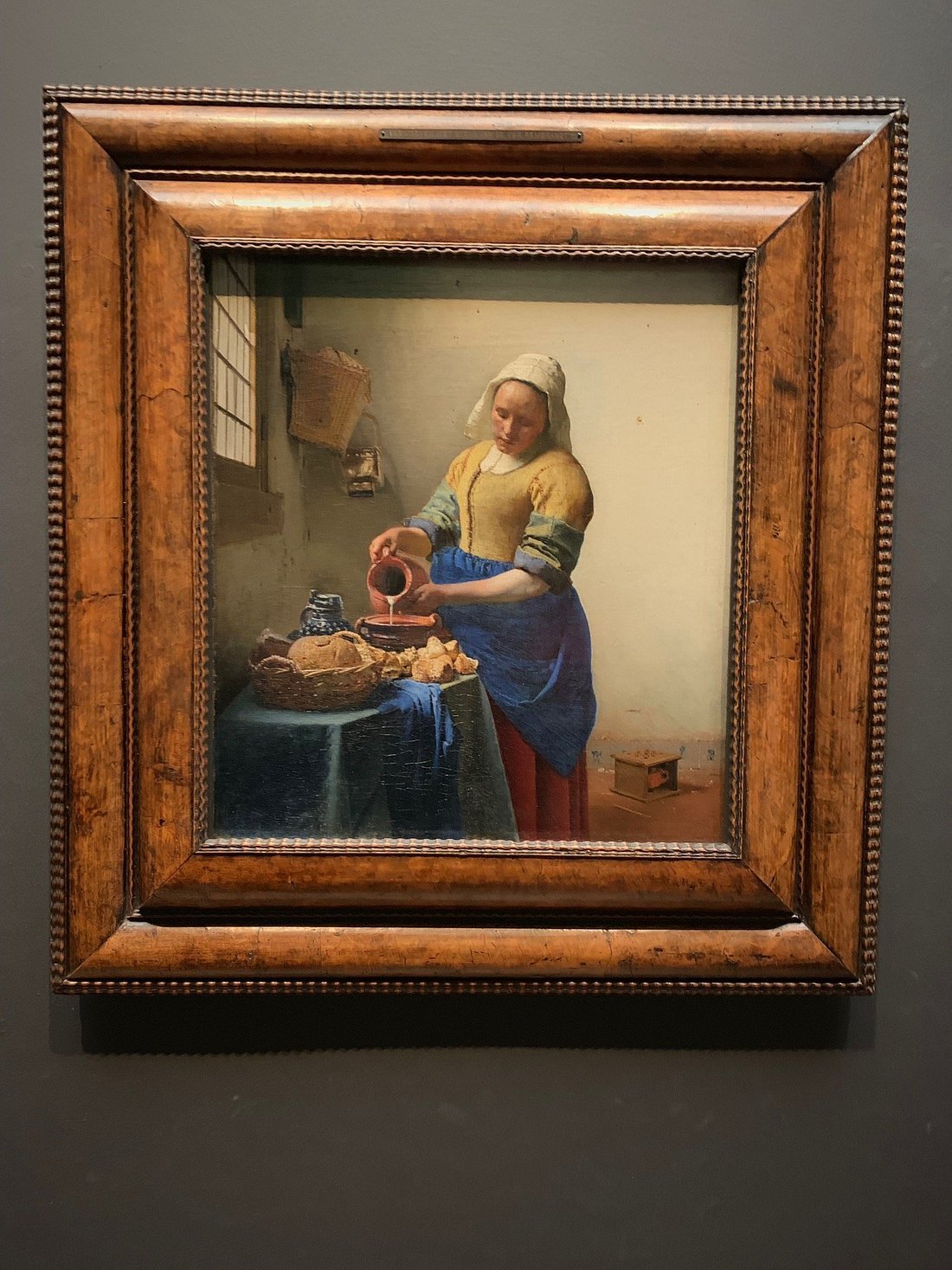 Vermeer's The Milkmaid in an ornate wooden frame on a dark gray wall at the Rijksmuseum.