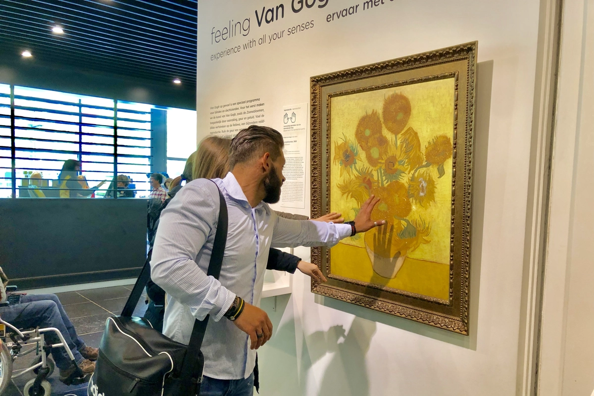 "Feeling Van Gogh" accessibility exhibit: a visitor touches a 3D relief of the Sunflowers painting in a gold frame.