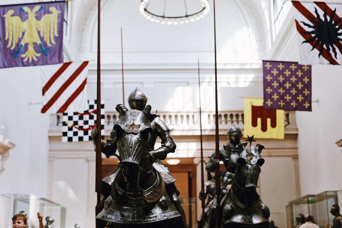 Medieval knights in plate armor on horseback displayed in the Met Museum's Arms and Armor gallery beneath heraldic banners.