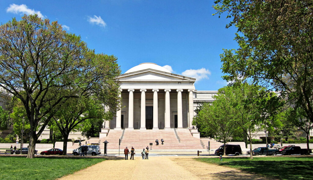 A neoclassical building with a dome and columns stands under a bright blue sky. Lush green trees frame the scene on either side. People and cars are visible in front of the building, and a wide pathway leads to the entrance.