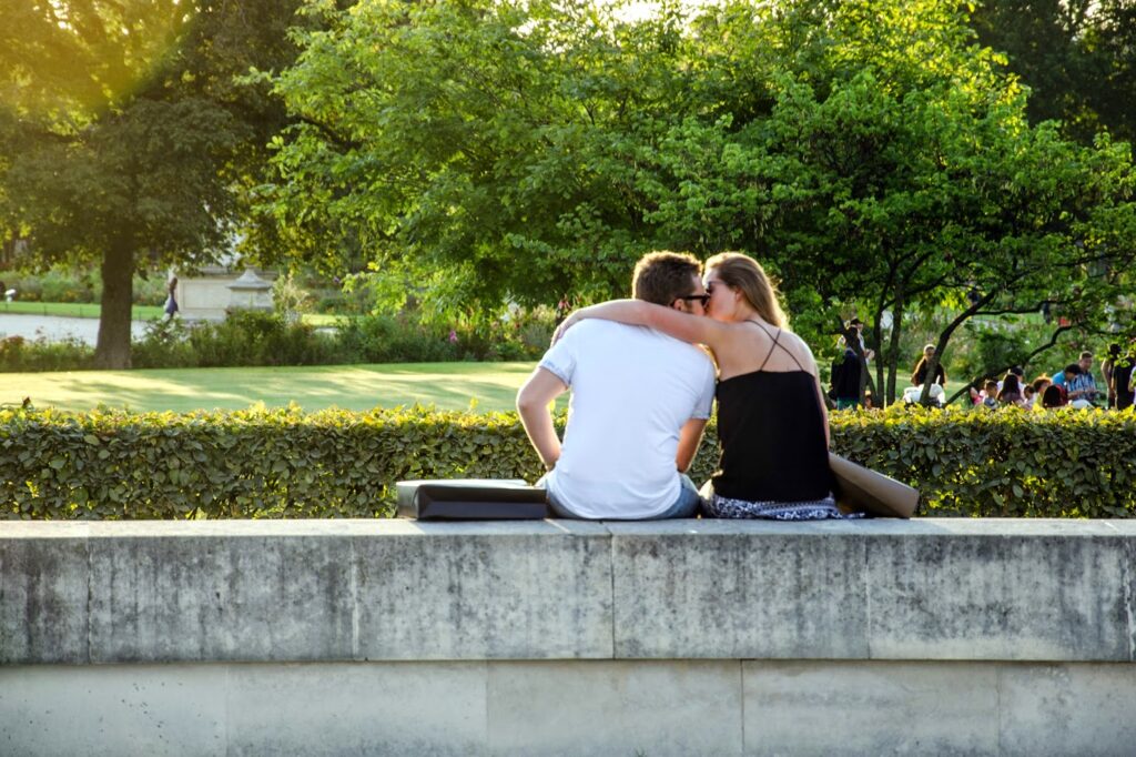 A couple sits closely on a stone ledge in a park, surrounded by greenery. The woman leans her head on the mans shoulder as they enjoy the sunny day. In the background, people stroll through the park and sunlight filters through the trees.