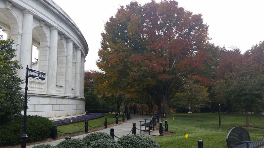 A curved white colonnade structure on the left, with a path lined by benches and lampposts leading through a park. A large tree with autumn foliage stands prominently, surrounded by green lawn and shrubs. Overcast sky in the background.