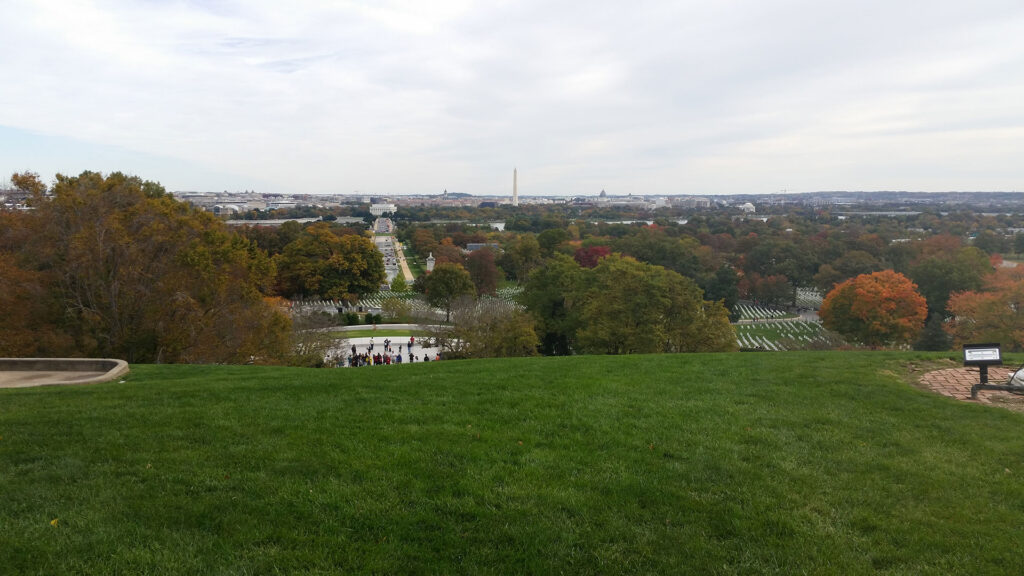 Scenic view from Arlington National Cemetery overlooking Washington, D.C. The landscape includes a lush green lawn, autumn trees, and distant monuments with a cloudy sky in the background.