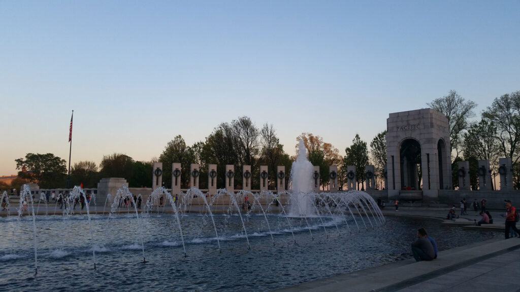 The image shows a large, oval-shaped fountain surrounded by a colonnade with stone pillars and an archway. Several people are seated nearby. An American flag is visible to the left, with trees in the background under a clear sky.