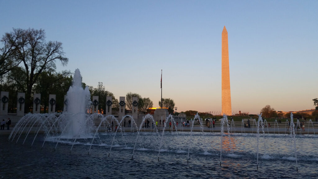 A sunset view of the Washington Monument, with its reflection in the foreground pool. Water fountains spray in the pool, and large trees and structures are silhouetted against the sky.