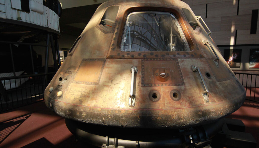 A close-up of a historic space capsule displayed in a museum. The metal exterior shows signs of wear, with visible windows and access panels. Subdued lighting creates shadows on the capsules surface.