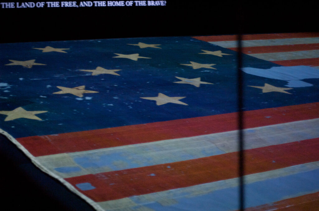 A partial view of a vintage American flag displayed in a dimly lit setting. The flag has stars and stripes, showing signs of wear and aging. A phrase from the national anthem is partially visible at the top.