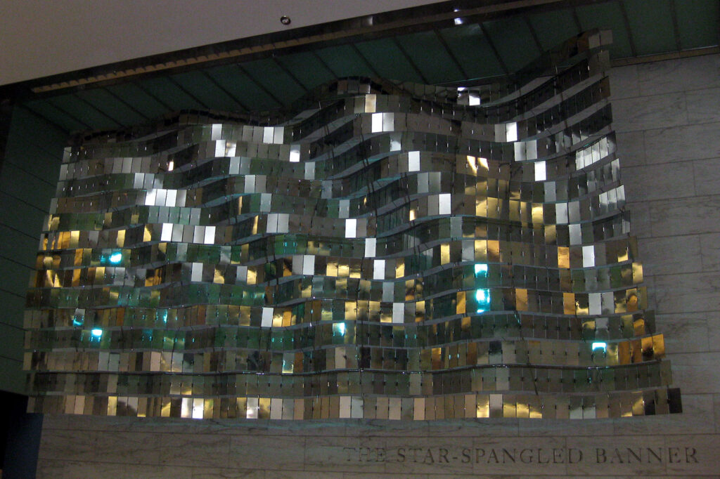 A wavy installation made of reflective metallic tiles, mounted on a wall. Beneath it, the text reads The Star-Spangled Banner. The tiles reflect ambient light, creating a shimmering effect.