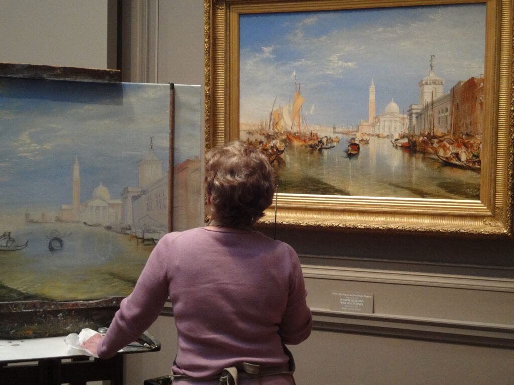 A person in a pink sweater works on a painting in a museum. The painting being worked on closely resembles the framed artwork on the wall, depicting a Venetian canal scene with boats and buildings under a blue sky.