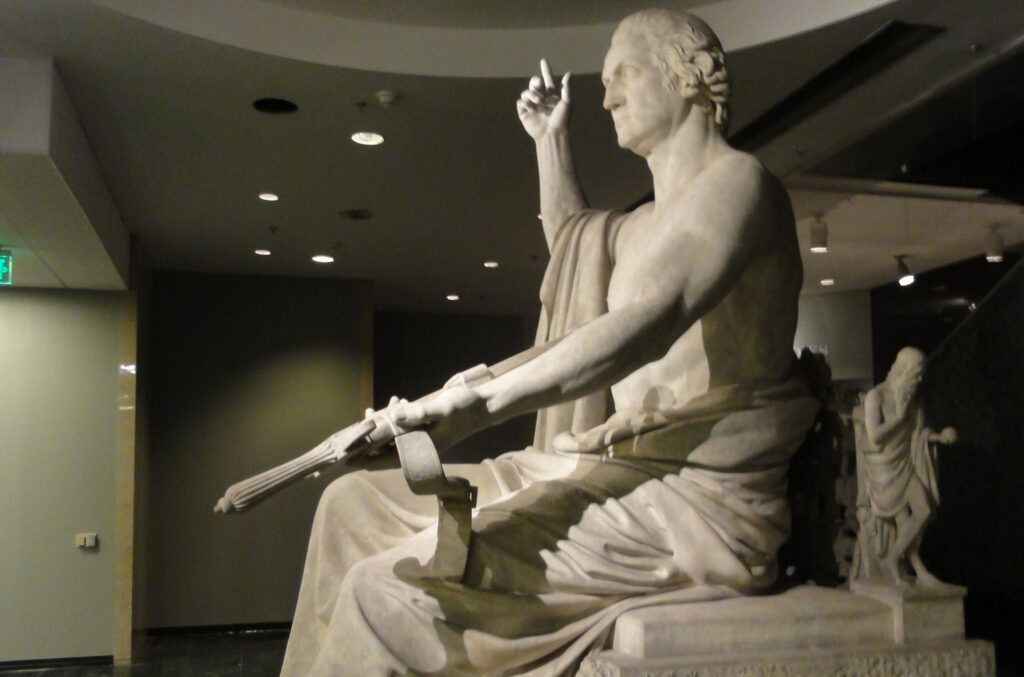 Large marble statue of a seated figure draped in classical robes, holding a staff. The figures right arm is raised, and it is set within a dimly lit room with a high ceiling.