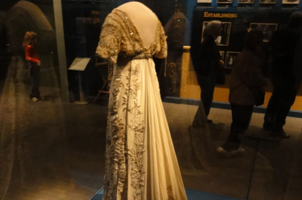 A vintage, ornate gown with intricate embroidery displayed in a museum exhibit. The dress features a long, flowing train and detailed beadwork on the bodice. Visitors are seen in the background observing the display.