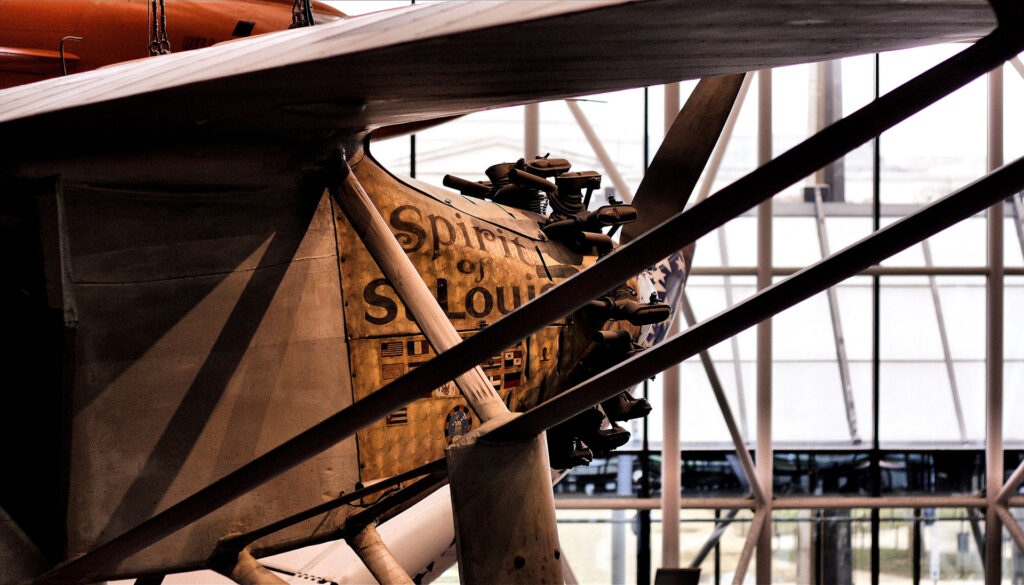 Close-up of an aircraft with Spirit of St. Louis painted on the side, showcasing its engine and structural components. The background features a grid of windows, allowing natural light into the space.