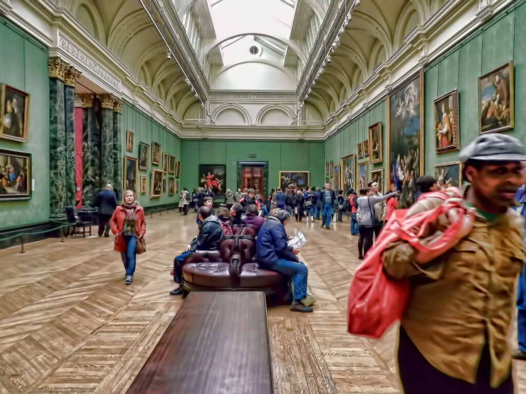 Visitors explore a large art gallery with high arched ceilings, green walls, and numerous paintings. People are seated on circular benches, while others walk around, observing the artwork.