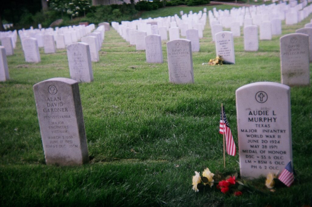 A serene cemetery scene with rows of white tombstones. The foreground shows a decorated grave with American flags and flowers. The grass is neatly trimmed, and trees are visible in the background.