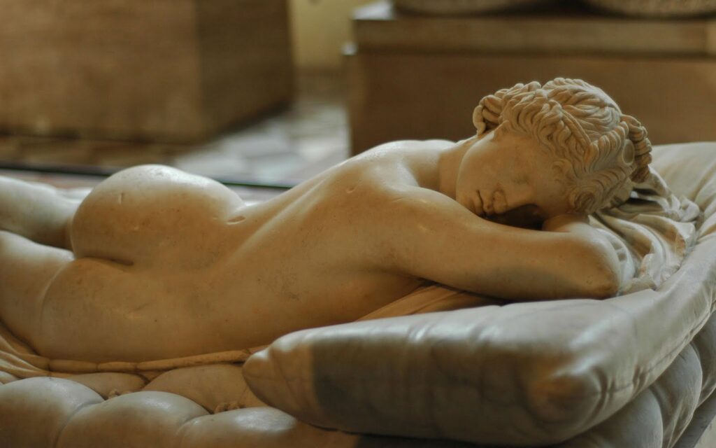 A marble statue depicts a reclining, nude figure lying face down on a cushioned surface. The detailed sculpture showcases the figures peaceful posture and intricate hair design. The background is blurred, highlighting the statues craftsmanship.