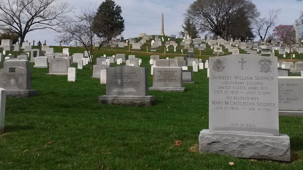 A cemetery with neatly arranged gravestones on green grass. The foreground features a gravestone with etched text. Trees and more gravestones are visible in the background, under a partly cloudy sky.