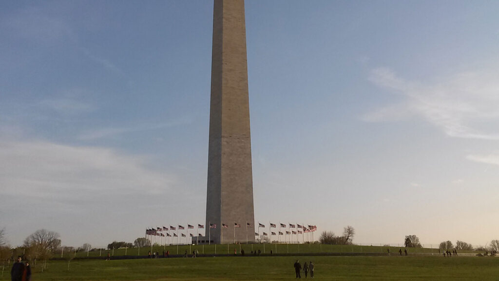 The Washington Monument stands tall against a clear blue sky. American flags circle the base, and people walk on the grassy area surrounding the monument.