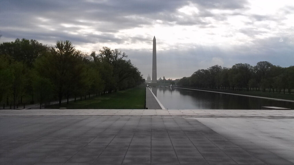 The image shows the Washington Monument reflecting in the Lincoln Memorial Reflecting Pool. The scene is framed by trees, with a cloudy sky allowing rays of sunlight to break through, creating a dramatic atmosphere.
