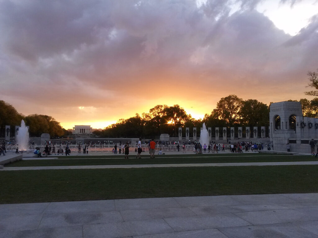 A sunset view over the World War II Memorial in Washington, D.C. The sky is a mix of vibrant orange and dark clouds. People are gathered around the reflective pool and fountains, with the Lincoln Memorial visible in the background.