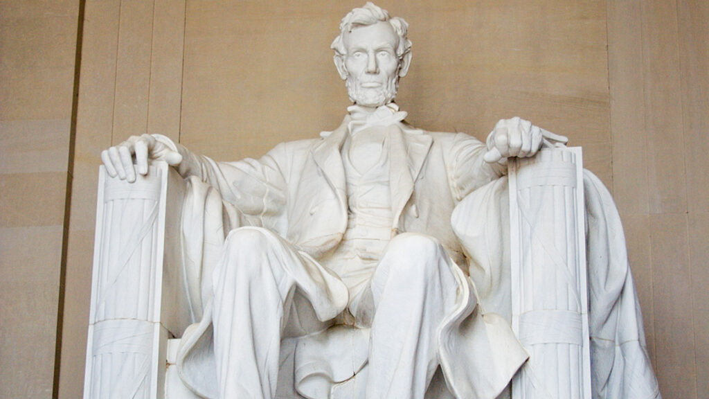 A statue of a seated man in a suit, with a contemplative expression, sitting on a large chair with arms resting on the sides. The statue is made of white marble and is set against a beige stone background.