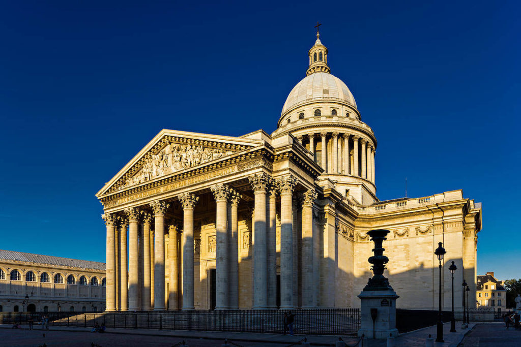 The image shows a neoclassical building with tall columns and a large dome, illuminated by sunlight against a clear blue sky. The building features intricate carvings above the entrance and cast shadows on the foreground.