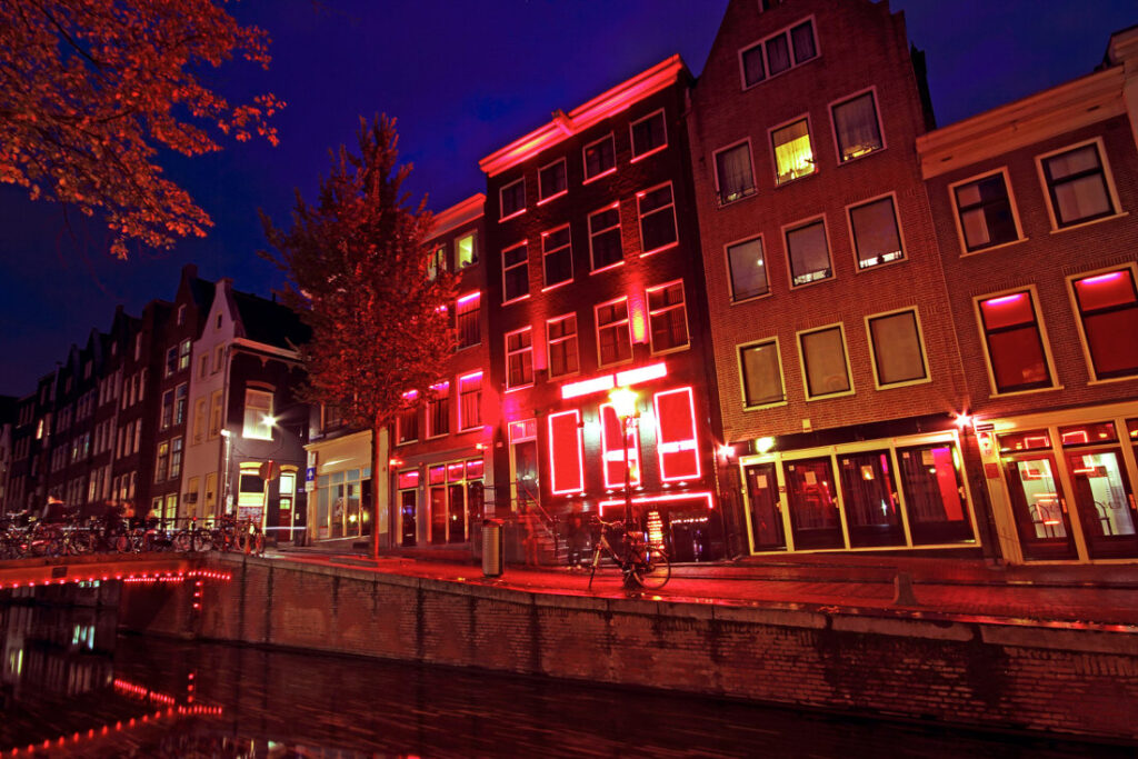 A canal in Amsterdam at night, featuring tall, narrow buildings with red lights reflecting on the water. Bicycles are parked along the canals edge, and trees add a touch of greenery to the urban scene.