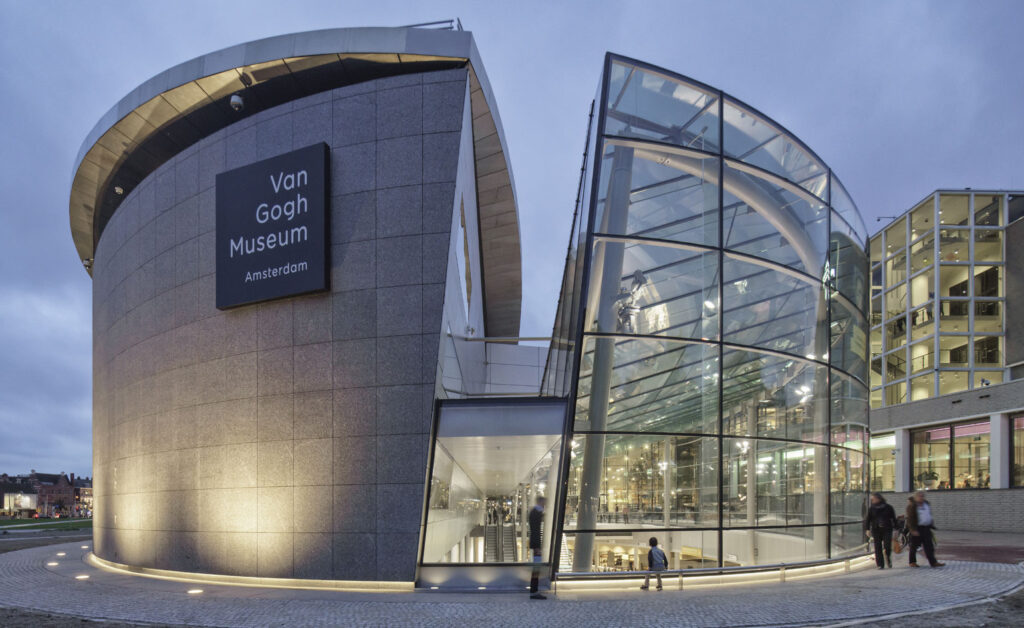 Modern entrance of the Van Gogh Museum in Amsterdam, featuring a curved glass facade and a sleek gray wall with the museums name. People are walking outside under a cloudy sky.