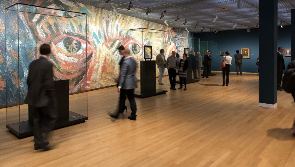 A museum gallery with people viewing artwork. The room has wooden floors, dark walls, and a large colorful abstract painting covering one wall. Several paintings are displayed in glass cases. Visitors are walking and observing the exhibits.