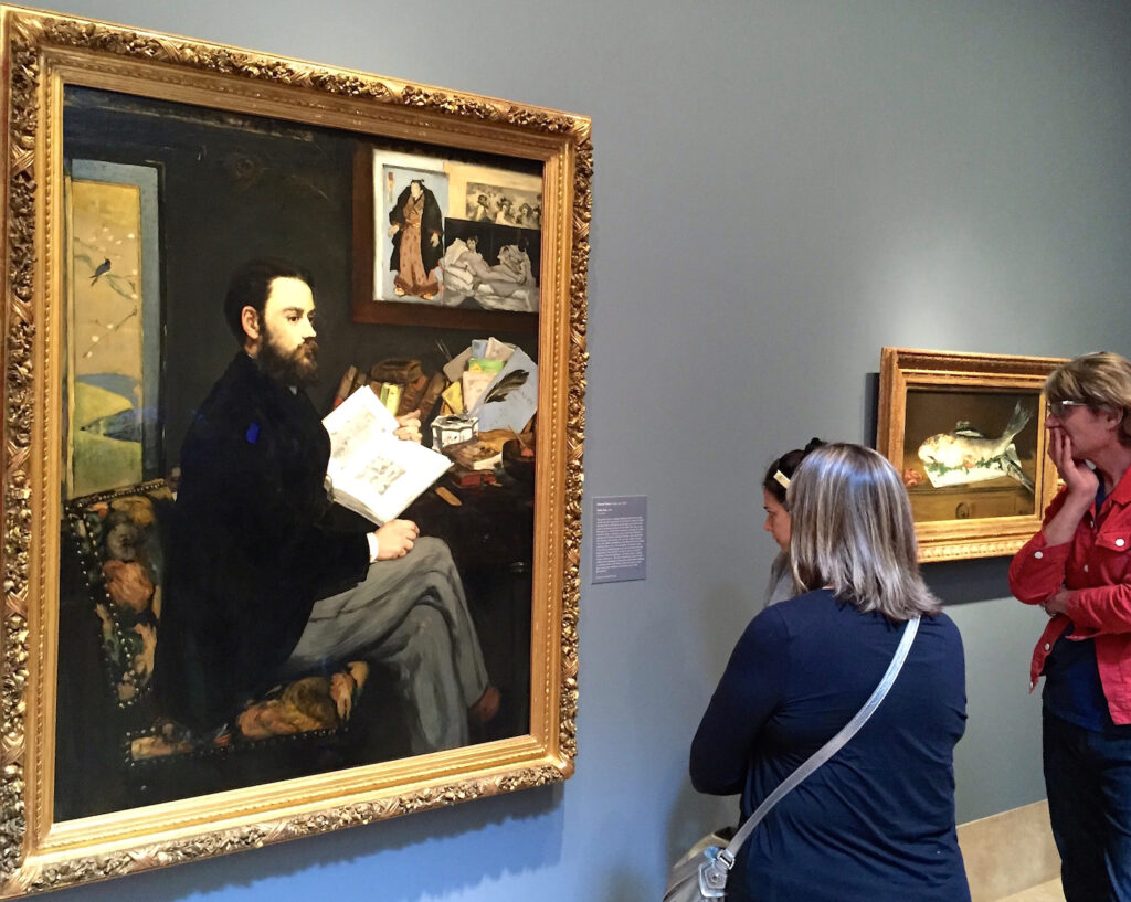 Three people observe a framed painting in a gallery. The artwork depicts a seated man with a book, surrounded by various items. Another smaller painting is visible on the wall to the right.