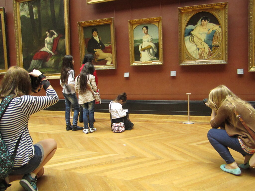 A group of people in a museum takes photographs and observes four classical paintings displayed on a red wall. Two individuals kneel for a closer look while capturing the artwork. The wooden flooring complements the gallery setting.