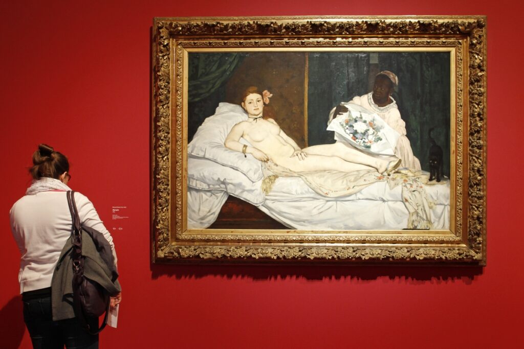 A woman looks at a framed painting of a reclining nude woman with a floral headpiece, attended by a servant holding a bouquet. The artwork contrasts against a vibrant red wall.