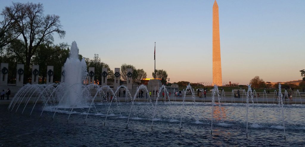 View of the Washington Monument with a reflecting pool and fountains in the foreground. The sun sets, casting a warm glow on the scene. Trees and a row of columns line the background, with a clear blue sky overhead.