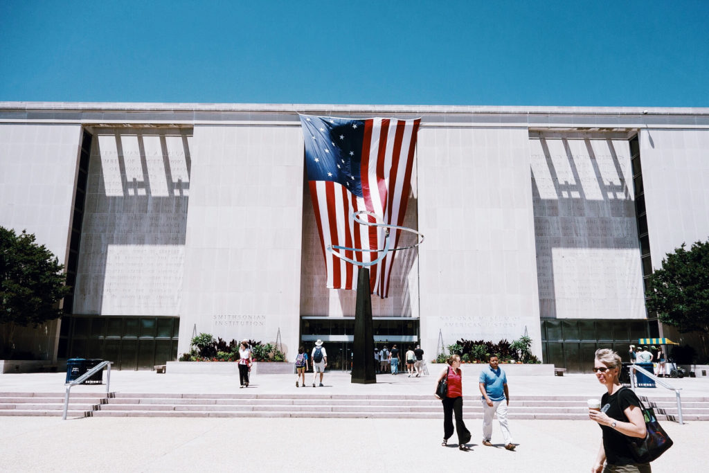 People walk in front of a large building adorned with a prominently displayed American flag sculpture. The structure has a modern design with steps leading to the entrance. The sky is clear and blue.