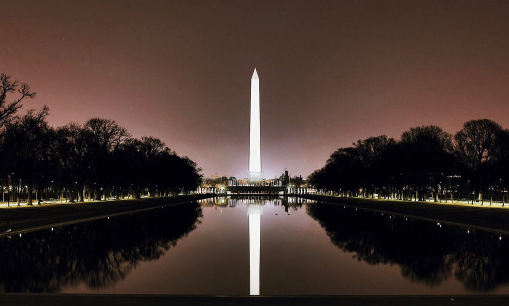 The image shows the Washington Monument at night, brilliantly lit and reflecting in the still waters of the Reflecting Pool. The scene is framed by leafless trees and a dark, clear sky, evoking a serene and majestic atmosphere.