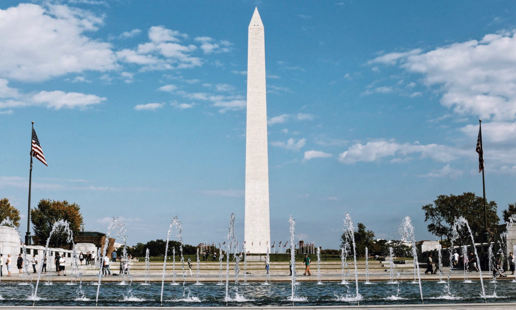 The image shows the Washington Monument against a blue sky with scattered clouds. In the foreground, water fountains are spraying, and American flags can be seen on poles on either side. Visitors are walking around the area.