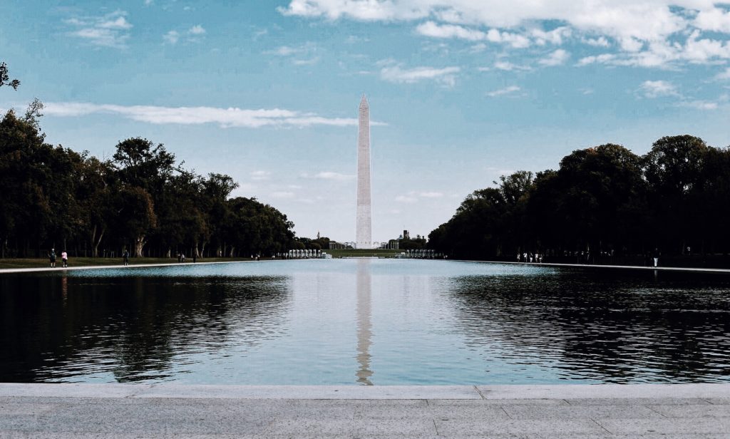 View of the Washington Monument with its reflection in the Reflecting Pool. The sky is partly cloudy, and the scene is framed by trees on both sides, creating a symmetrical composition.