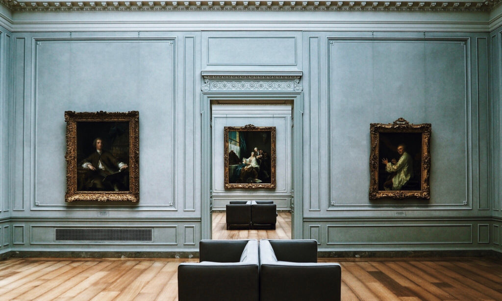 A spacious art gallery with three framed paintings on light blue walls. Two sofas are placed on a wooden floor, offering a view of the artworks. The gallery has intricate molding details on the ceiling and walls.