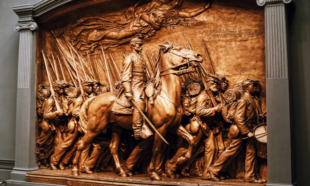 A detailed bronze relief sculpture depicting a group of soldiers marching, led by a figure on horseback. The soldiers carry rifles and wear uniforms, with flags waving in the background, conveying a sense of movement and historical significance.