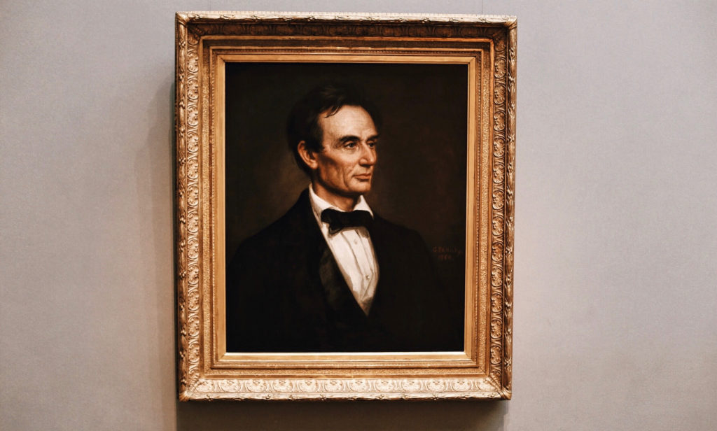 A framed painting of a man in a dark suit and bow tie, set against a dark background. The frame is ornate with intricate detailing. The man has a thoughtful expression and the painting is displayed on a neutral-colored wall.