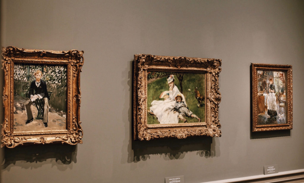 Three framed paintings hang on a gray wall in a gallery. Each artwork features figures in various settings, surrounded by ornate gold frames. The lighting highlights the details and colors of the paintings.