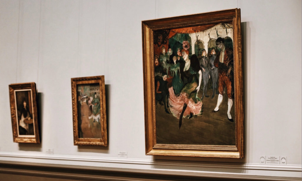 A gallery wall featuring three framed paintings. The largest painting depicts a lively scene with figures in period clothing. The two smaller paintings on the left show more subdued figures in intimate settings. The wall is painted a neutral color.
