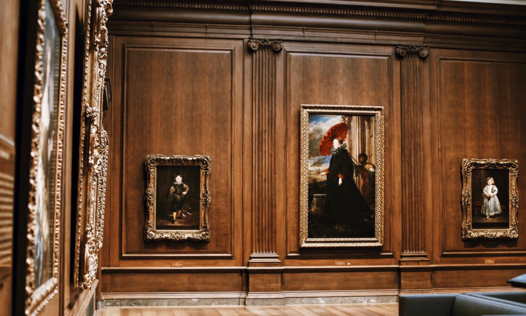A museum gallery featuring several framed paintings on ornately decorated wooden walls. The central painting depicts a figure with a red umbrella. The floor is wooden, and a bench is partially visible at the bottom right.