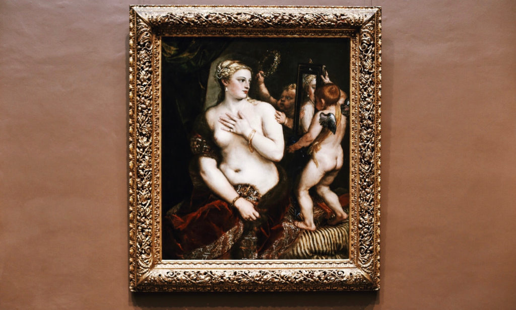A classic painting in an ornate gold frame depicts a woman gazing into a mirror held by a cherub, showcasing elaborate details and rich colors on a plain tan wall.
