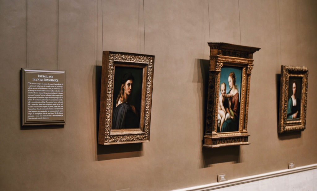 A museum wall displays three framed Renaissance paintings. The first frame shows a portrait of a man, the second a Madonna and Child scene, and the third another portrait. A descriptive plaque is mounted on the left wall.