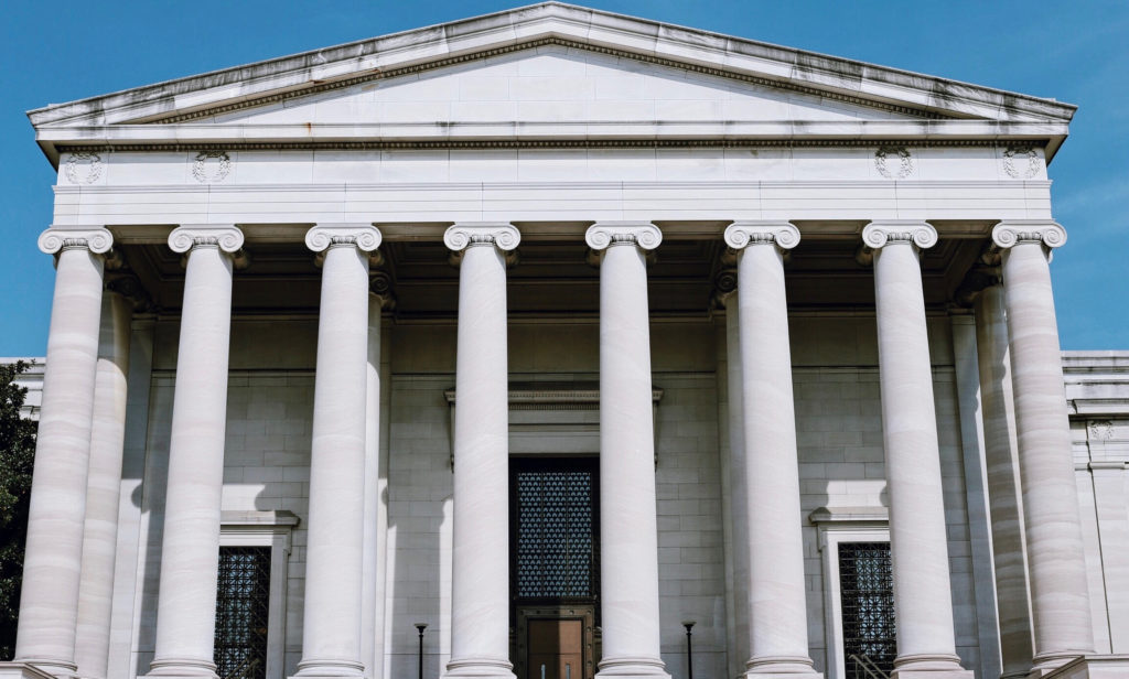 A classical Greek-style building with a large white facade and six tall columns under a triangular pediment. The clear blue sky serves as the backdrop.